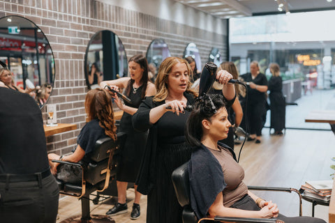 Woman getting her hair styled in a salon with other staff and customers in the background.